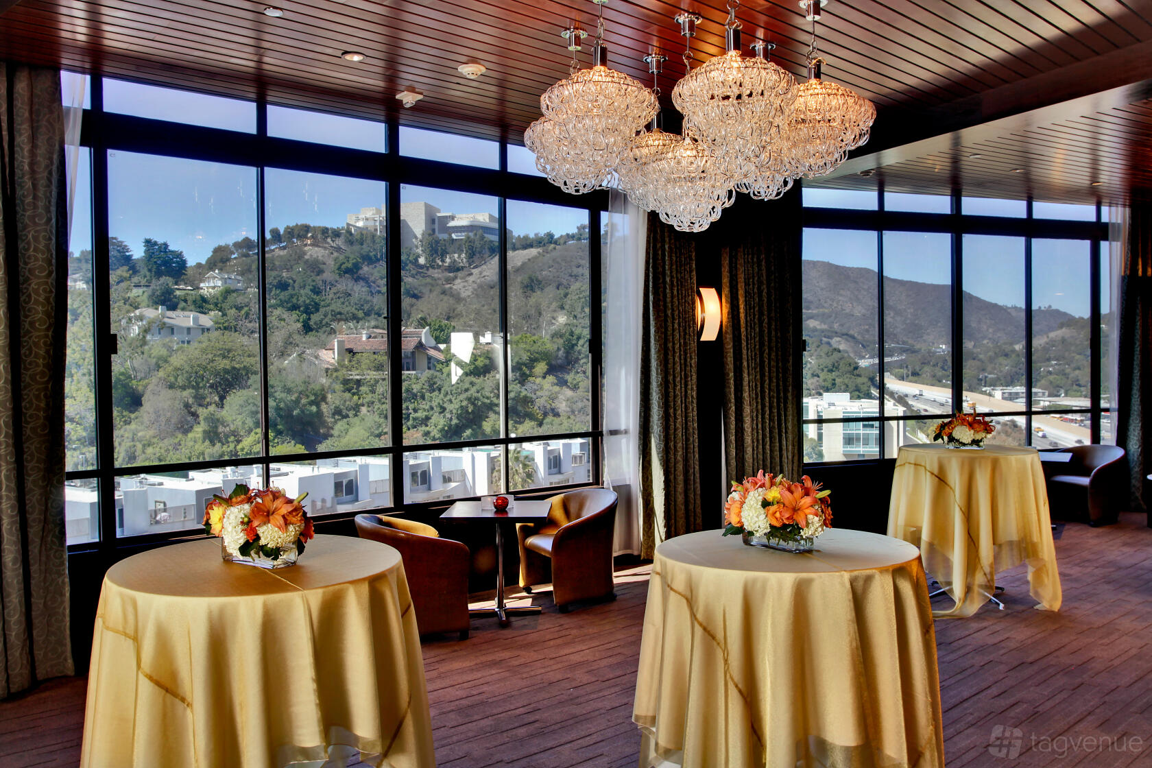 A function room with floor-to-ceiling windows, round cocktail tables with gold linens, and chandeliers at Hotel Angeleno.