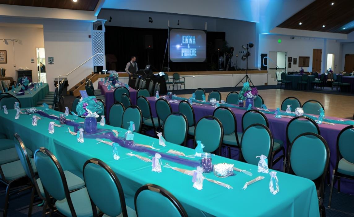 An event hall with teal round-backed chairs, long tables covered in turquoise linens, and overhead stage lighting at Adat Shalom Banquet Hall.