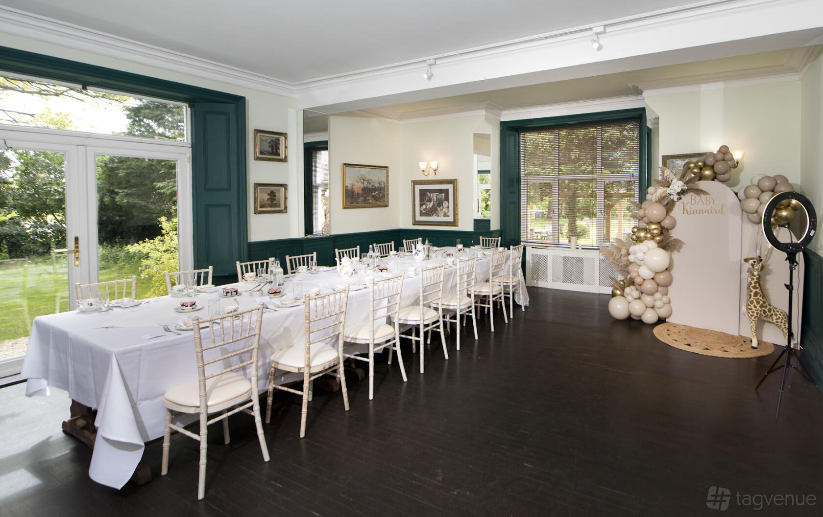 A dining room with a long table covered in white linens, chiavari chairs, and floor-to-ceiling windows at Trunkwell House Hotel.