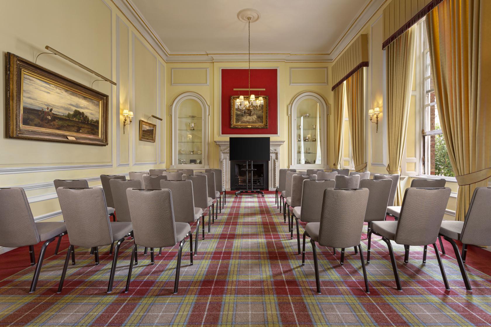 An event venue with tartan carpet, rows of cushioned chairs, tall windows, and classical wall art at The Caledonian Club.
