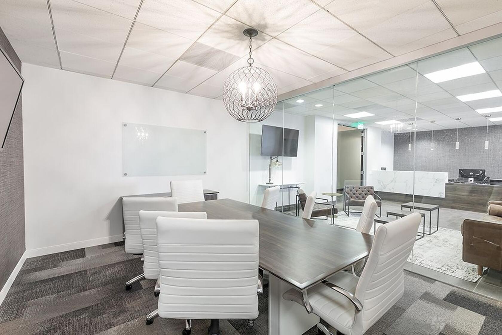 A coworking space with a glass wall conference room, white leather chairs, and a modern chandelier at Premier Workspaces - Wells Fargo Center.