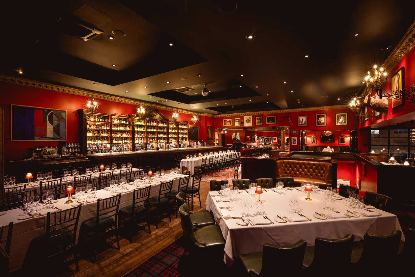 A restaurant with candlelit long tables, red walls, framed art, and a stocked backlit bar at Boisdale of Canary Wharf.