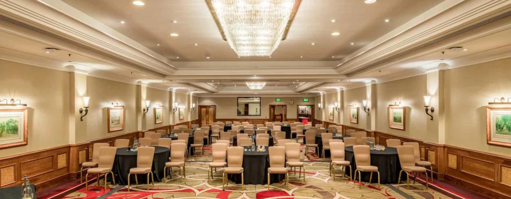 A hotel hall with rows of beige chairs, black-covered tables, patterned carpet, and a large crystal chandelier at The Marble Arch Hotel.