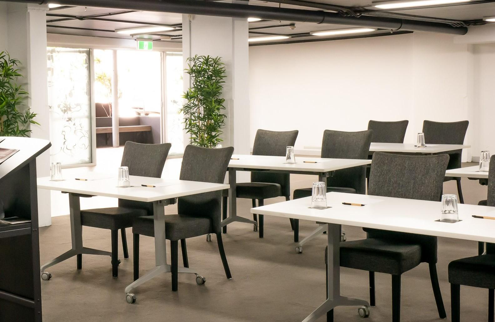 A hotel meeting room with rows of gray chairs, white rectangular tables, and potted plants at Punthill Apartment Hotel Spring Hill.