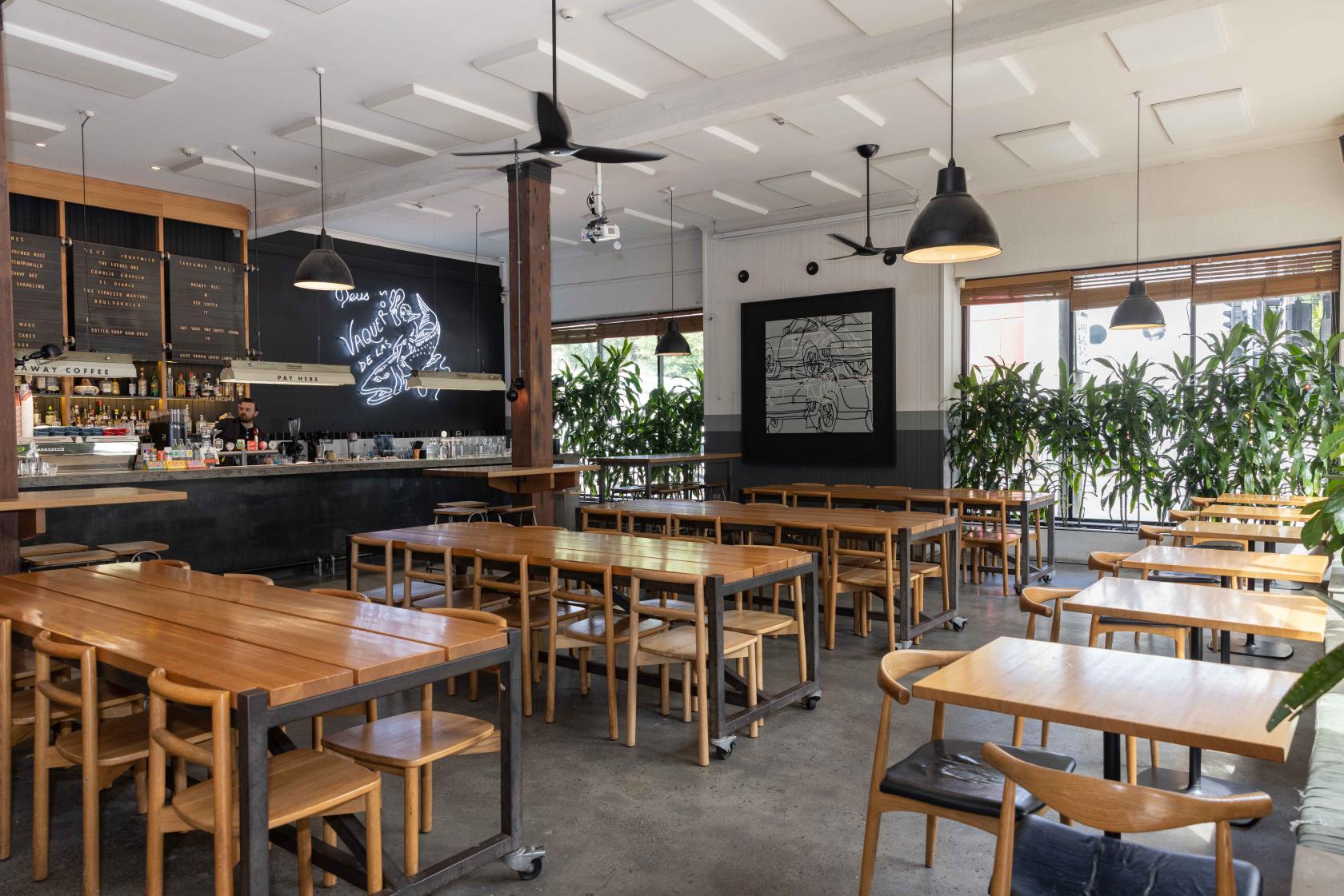 A cafe with long wooden tables, pendant lighting, and large windows with potted plants at Deus Cafe.