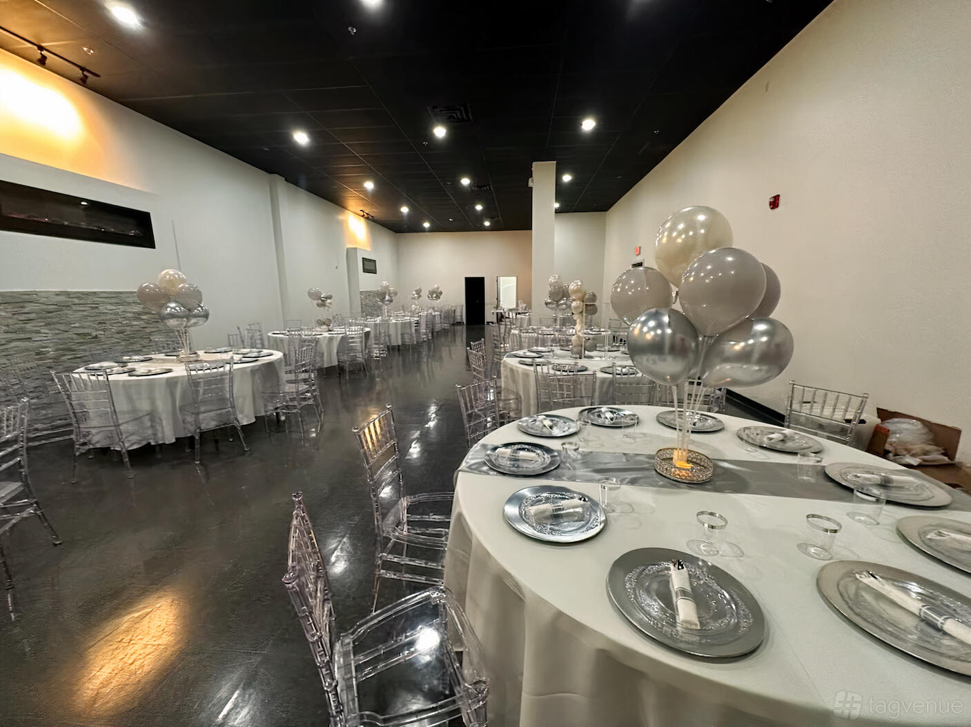 An event venue with round tables dressed in white linens, silver balloons, and clear chairs at Grand Luxe Vue.
