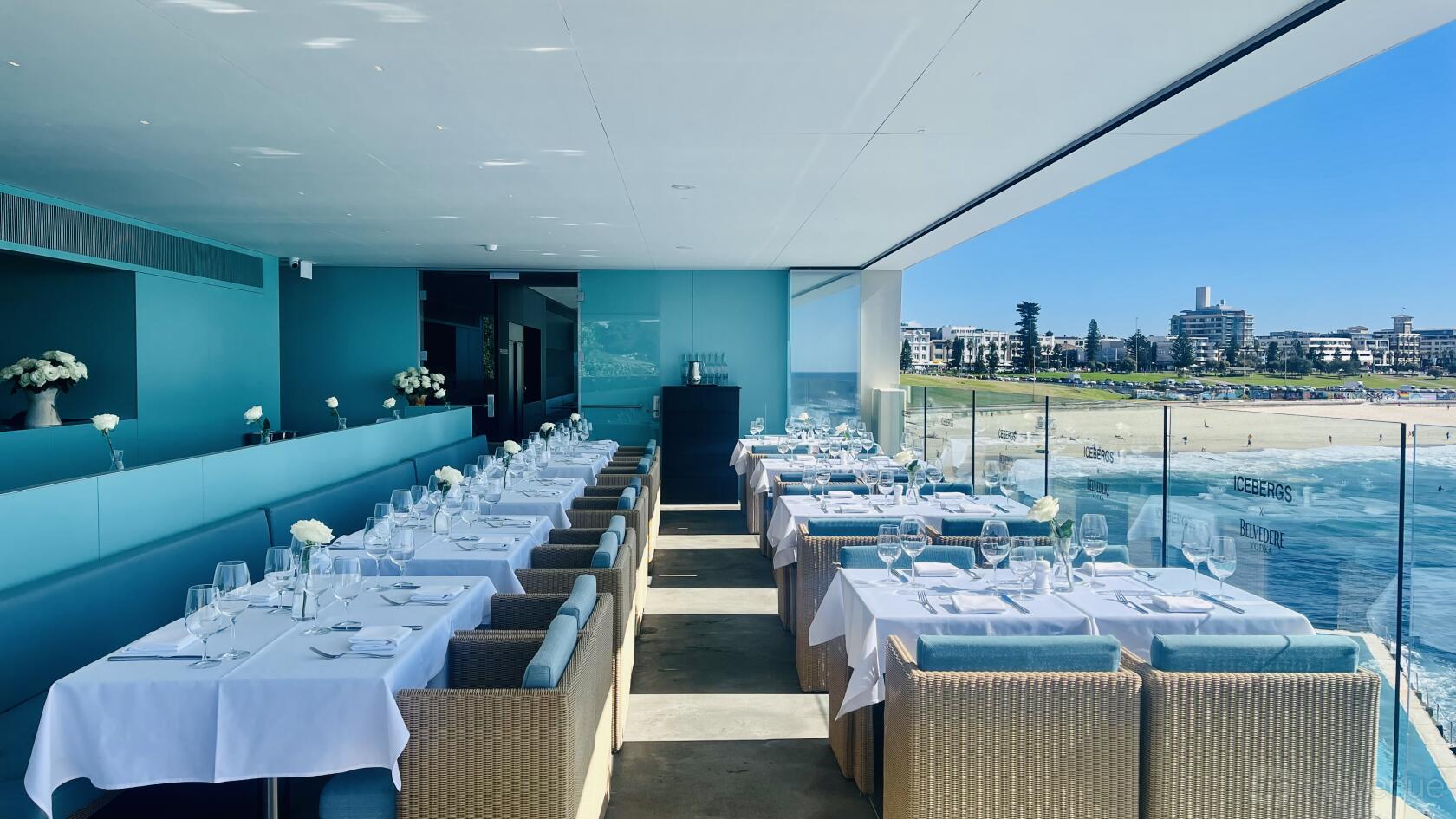 A function room with white linen tables, ocean views through glass walls, and blue accents at Icebergs Dining Room.