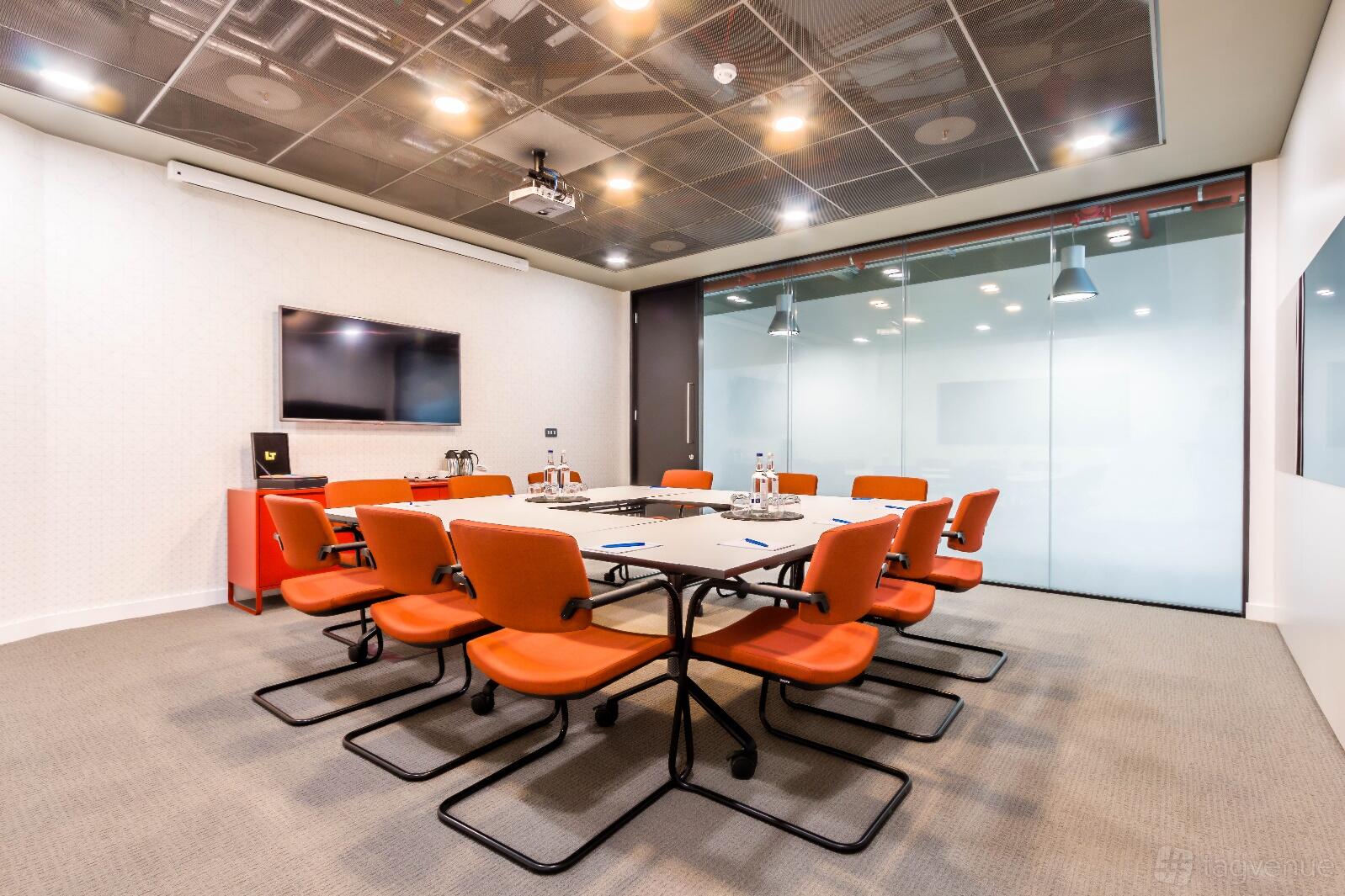 A meeting room with a large conference table, orange chairs, wall-mounted screen, and glass partition at Paddington.