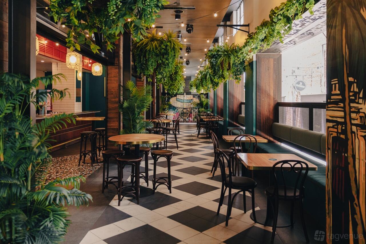 A bar with hanging greenery, checkered tile floors, round wooden tables, and black chairs at Hey Chica.