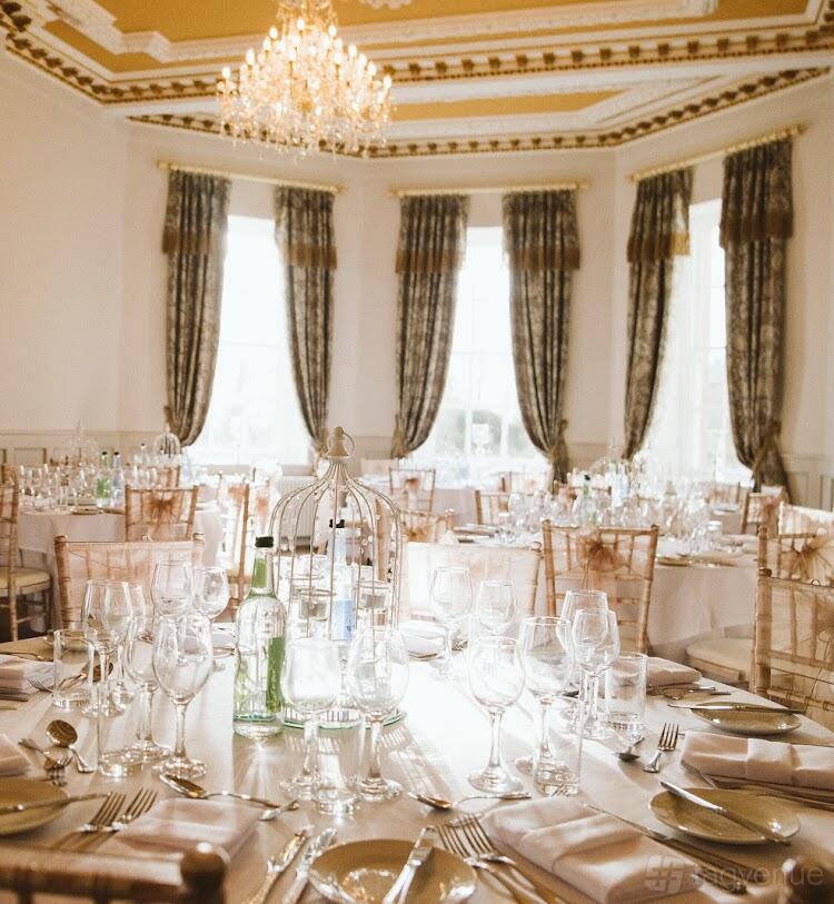A banquet hall with tall windows, elegant drapes, and round tables set with glassware at Bourton Hall.