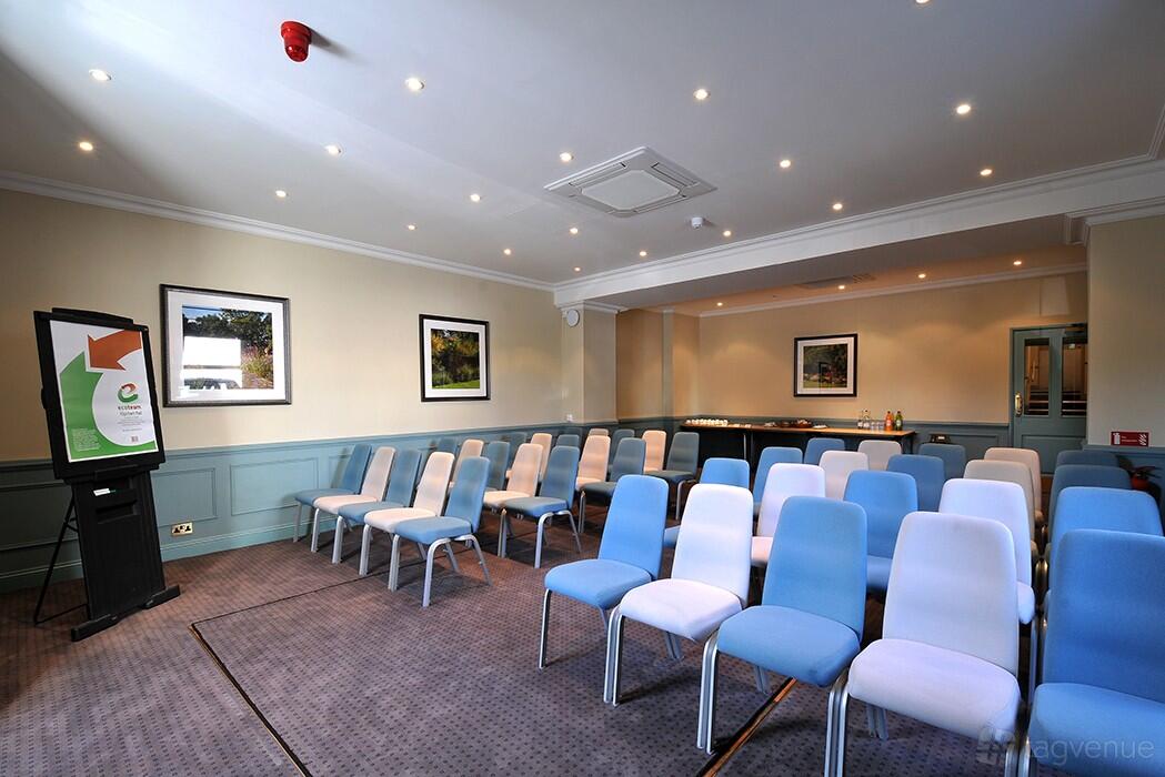 A meeting room with rows of blue and white chairs, carpeted floor, and framed wall art at The Bromley Court Hotel.