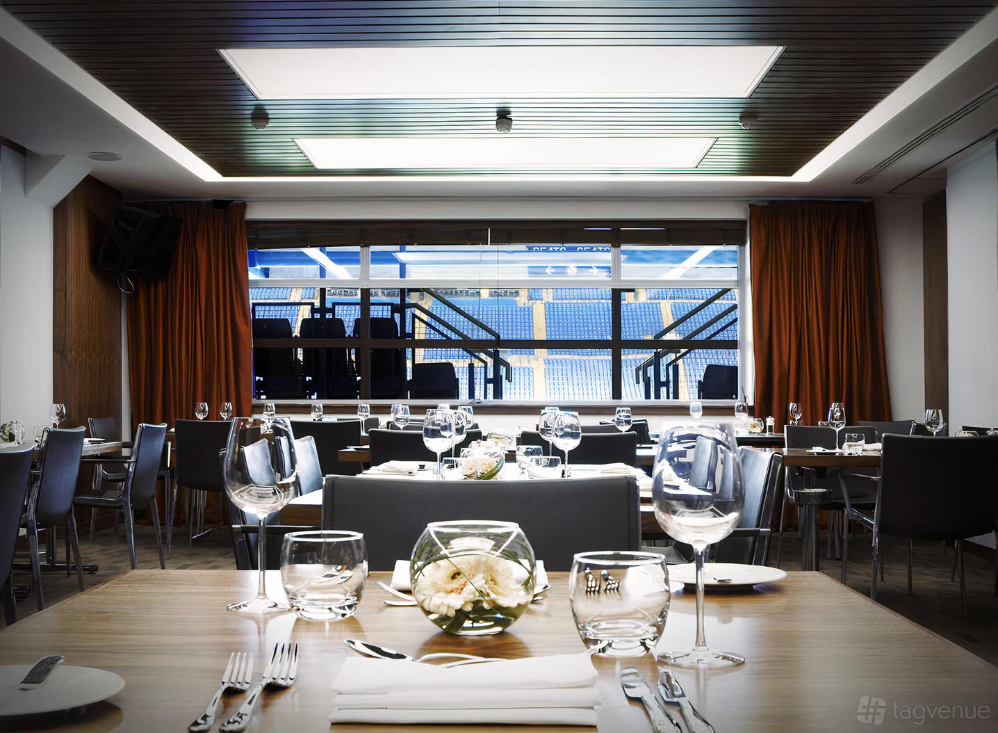 A stadium suite with floor-to-ceiling windows, set dining tables, and views of the pitch at Chelsea Football Club.