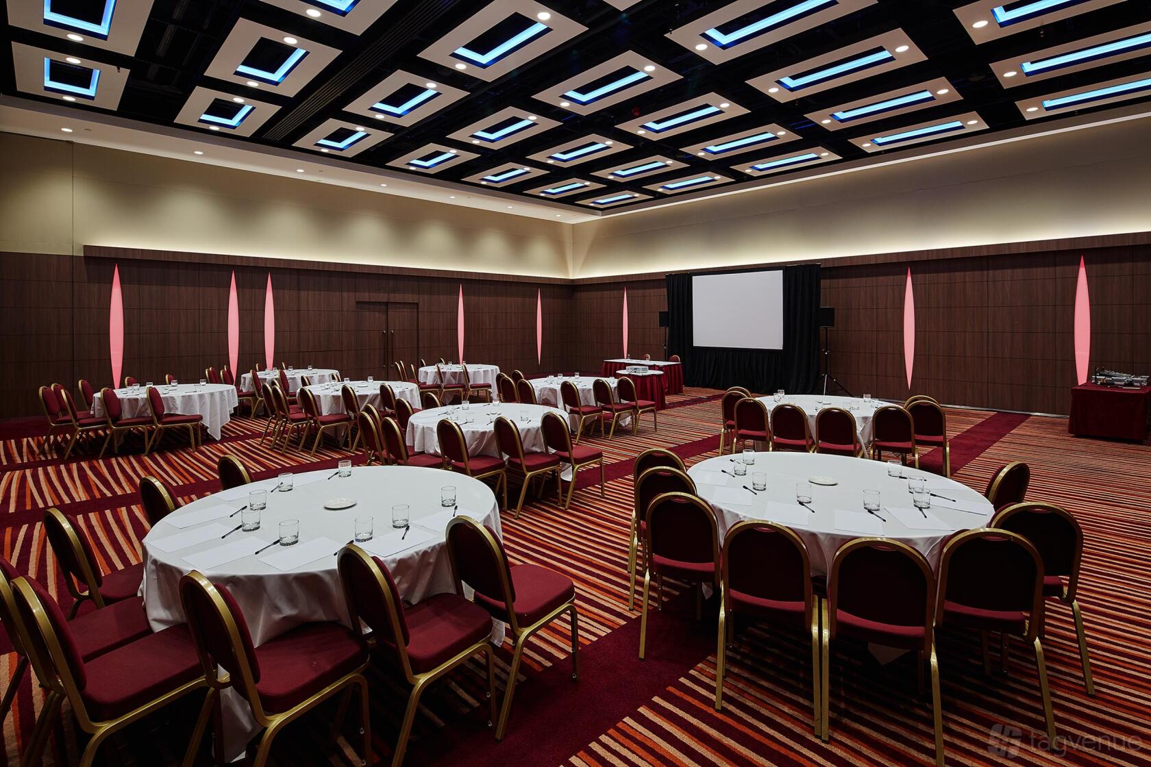 A hotel meeting room with round tables, red chairs, striped carpet, and a large projector screen at Mercure London Earl's Court.