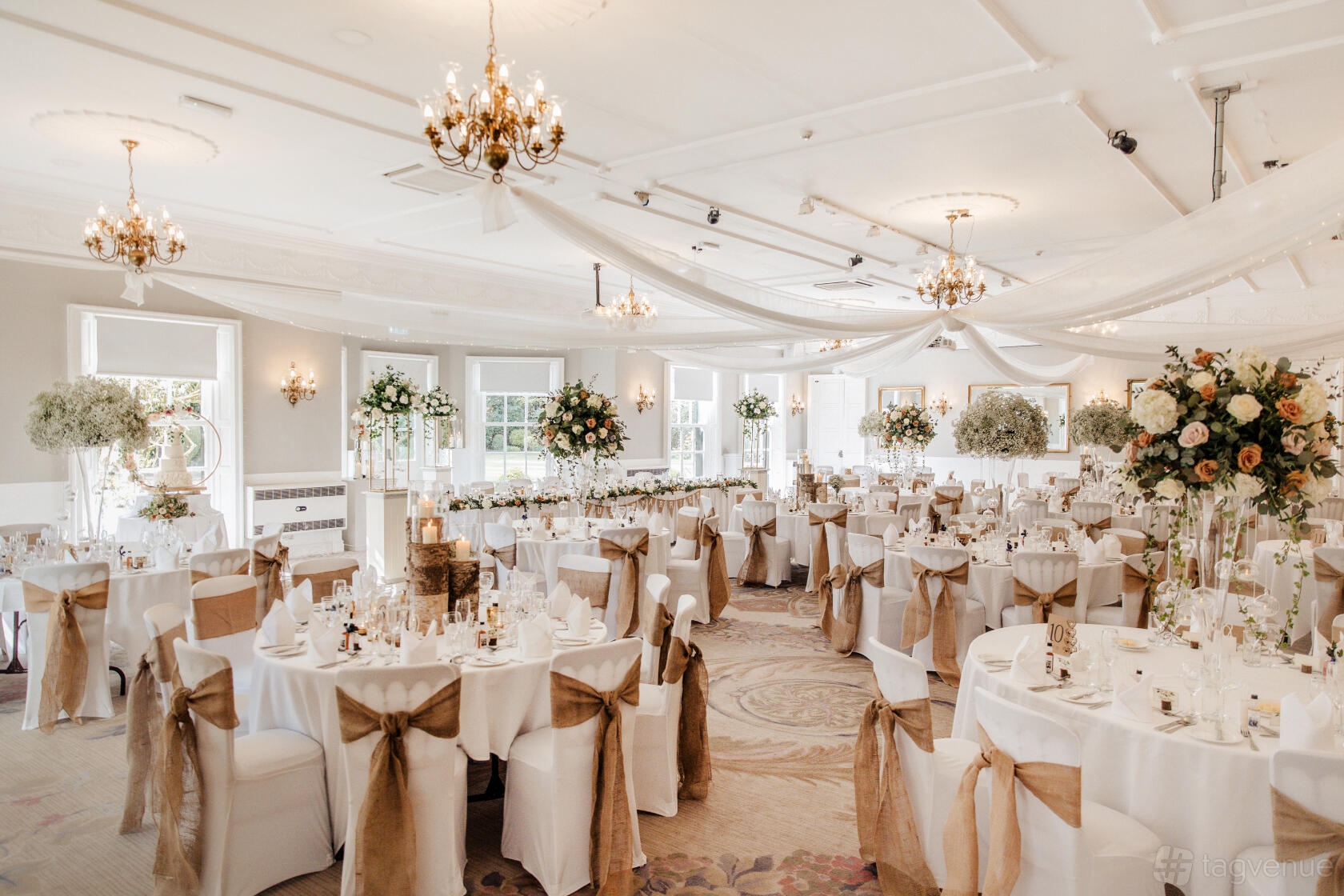 An event space with round tables draped in white linens, floral centerpieces, and chandeliers at Statham Lodge Hotel.