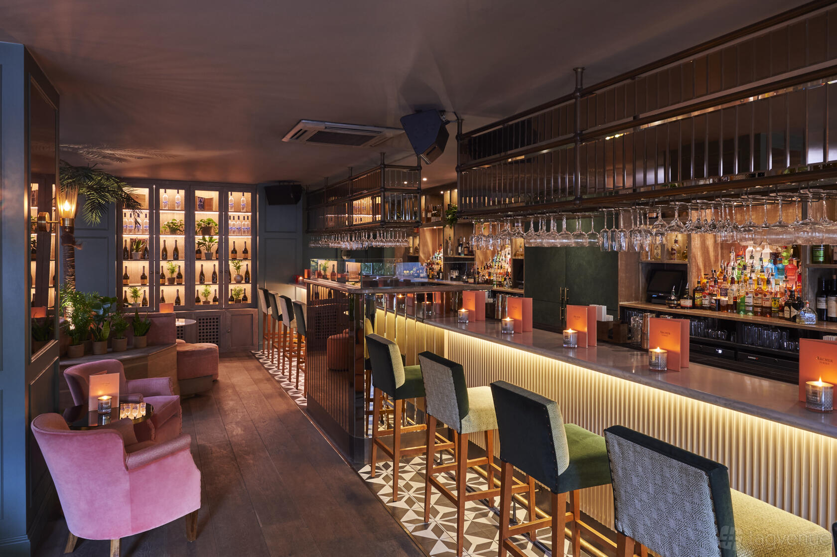 A bar with illuminated shelving, velvet armchairs, and a long counter with high stools at Archer Street Notting Hill.
