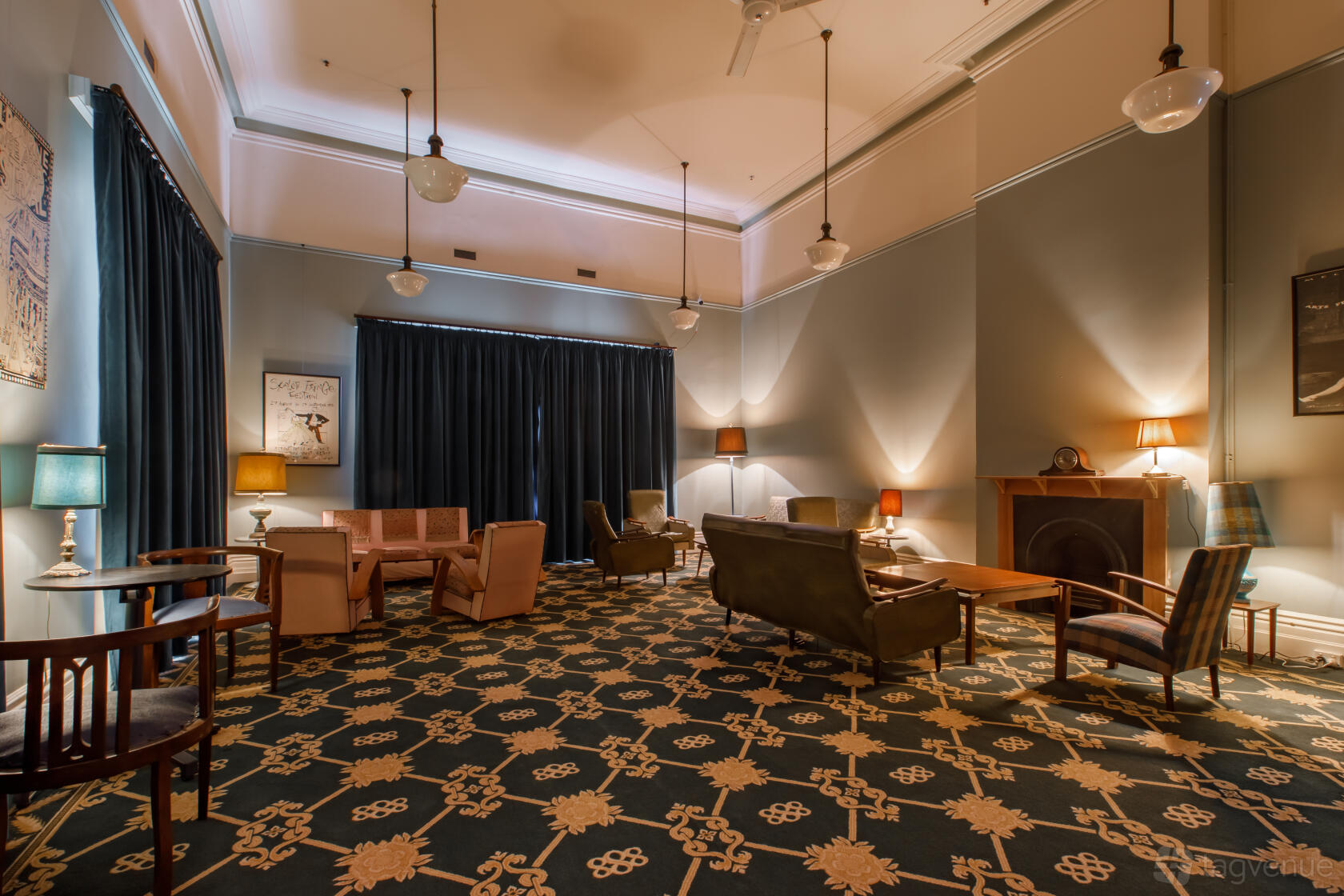 A lounge with patterned carpet, vintage armchairs, floor lamps, and a fireplace at Fringe Common Rooms.