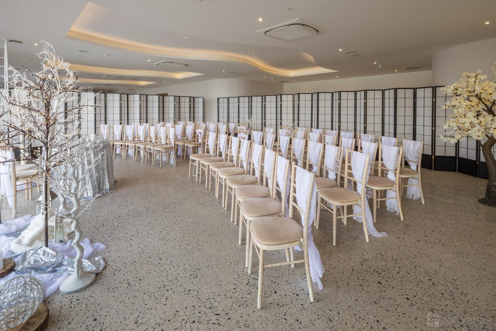 A hotel event space with beige chairs draped in white fabric, decorative trees, and terrazzo floors at The Venue At Sandy Cove.