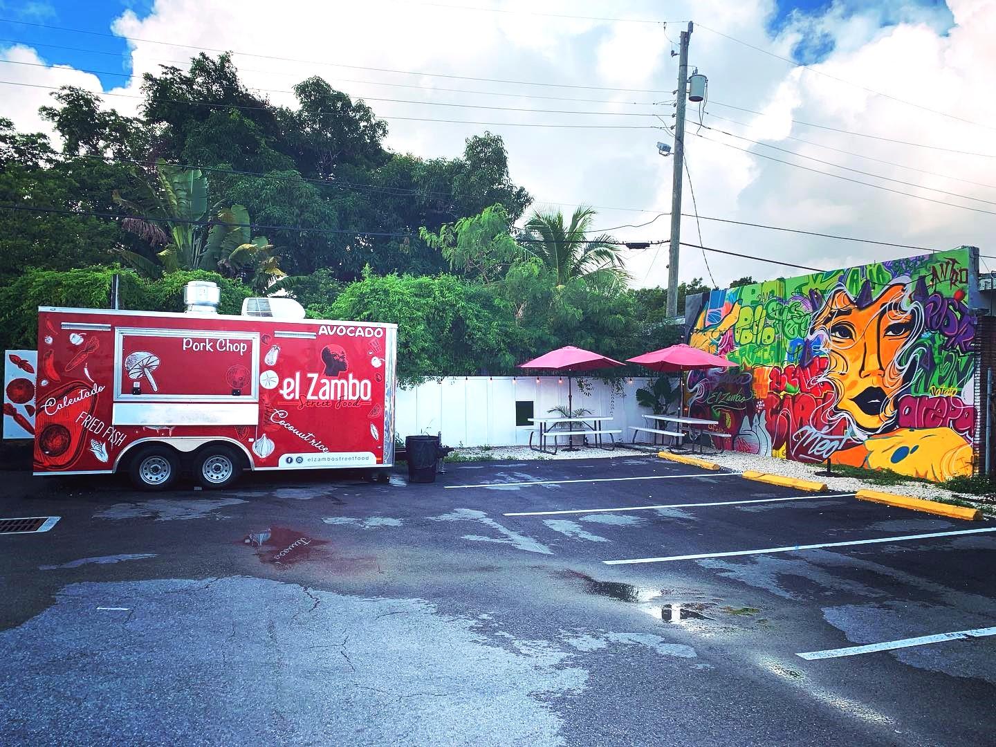 An outdoor restaurant space with a red food truck, street art mural, and picnic tables with umbrellas at El Zambo Street Food.