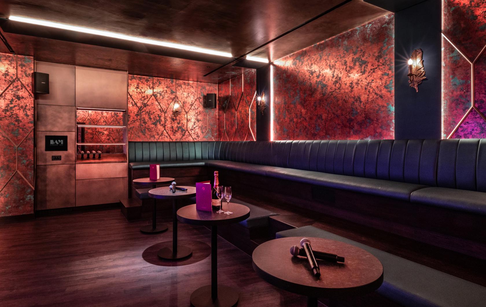 A function room with plush blue banquette seating, red textured walls, round tables, and ambient lighting at BAM Karaoke Box.