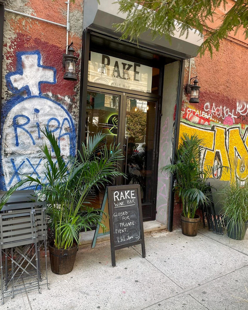 An entrance to a bar with graffiti-covered walls, potted plants, and a chalkboard sign at Rake Wine Bar.