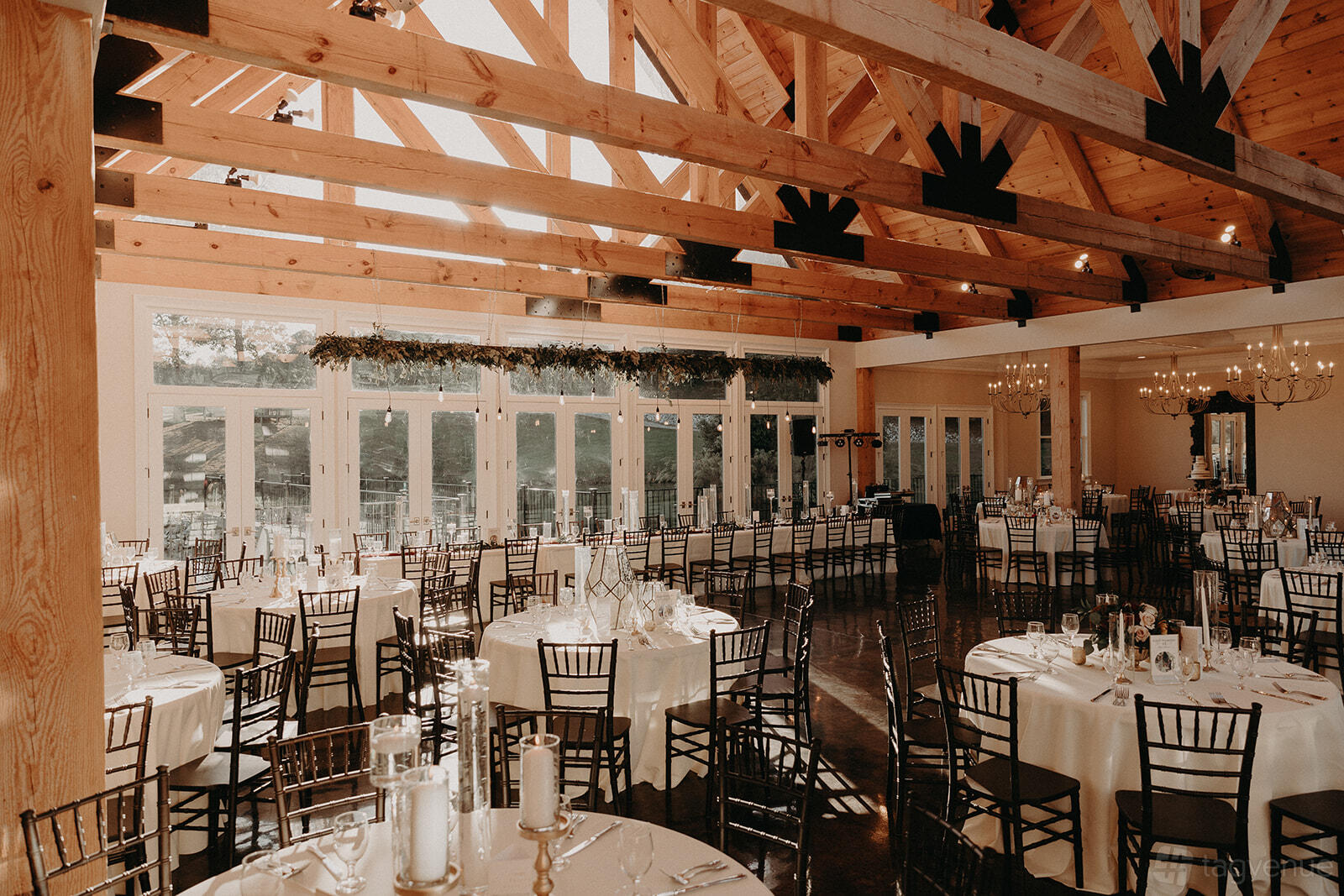 An event venue with exposed wooden beams, round tables with white linens, and floor-to-ceiling windows at Swan Lake Overlook.