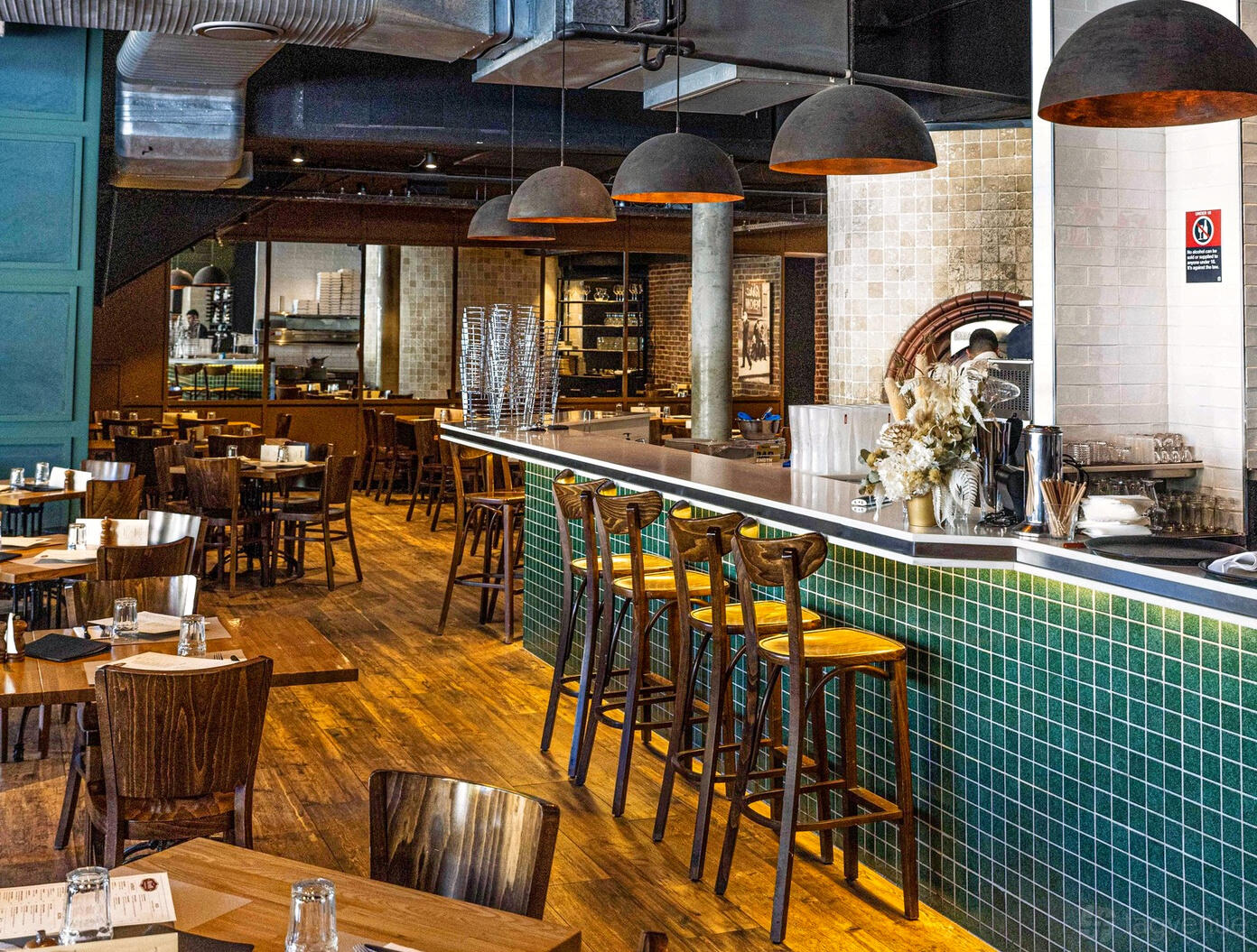 A restaurant with wooden tables, green tiled bar, hanging black pendant lights, and open kitchen at Moretti Ristorante Pizzeria.