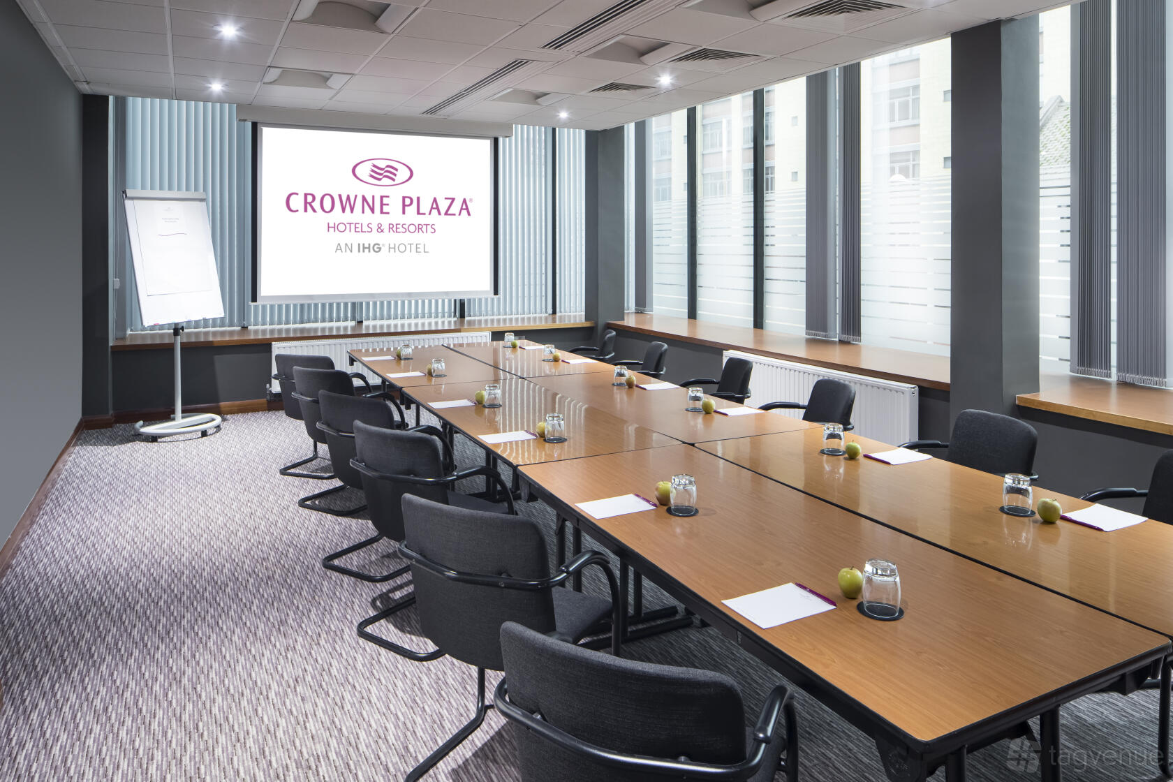 A boardroom with a U-shaped table, black chairs, a projector screen, and large windows at Crowne Plaza Nottingham.