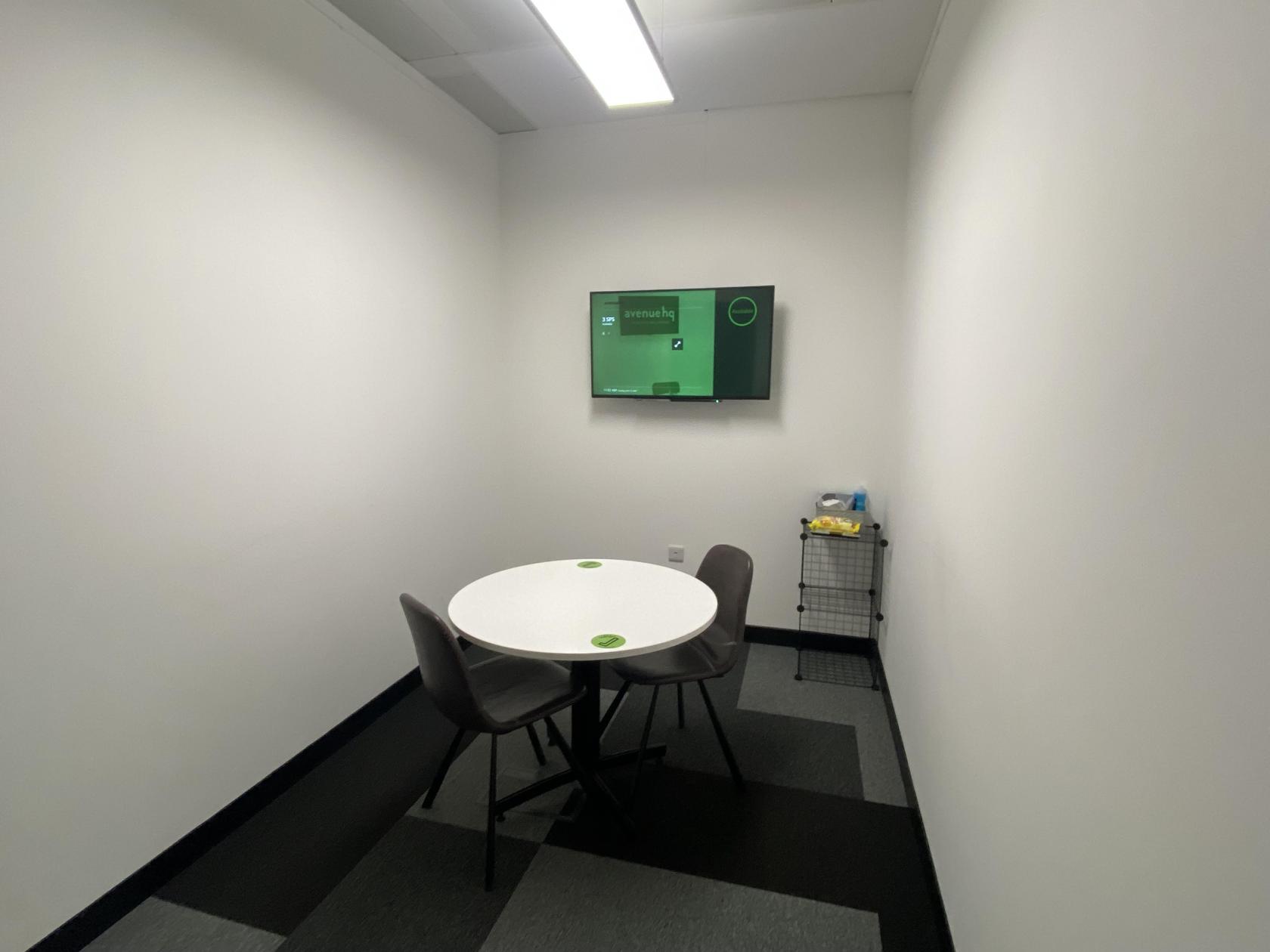 A coworking space with a round table, three chairs, white walls, and a wall-mounted screen at Avenue HQ St Paul's Square.