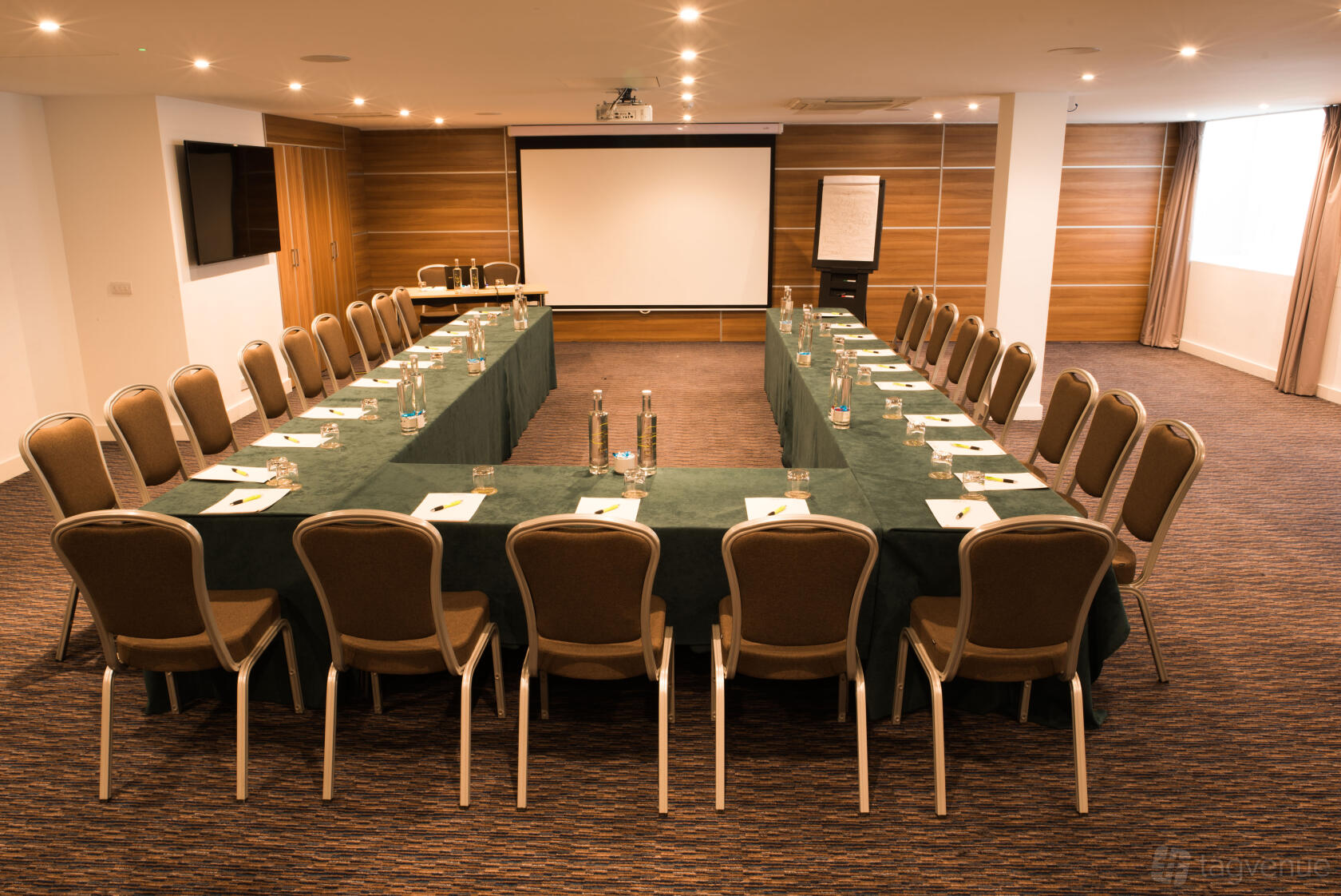 A function room with U-shaped tables, notepads, water bottles, and a projector screen at The Wesley Euston Hotel.