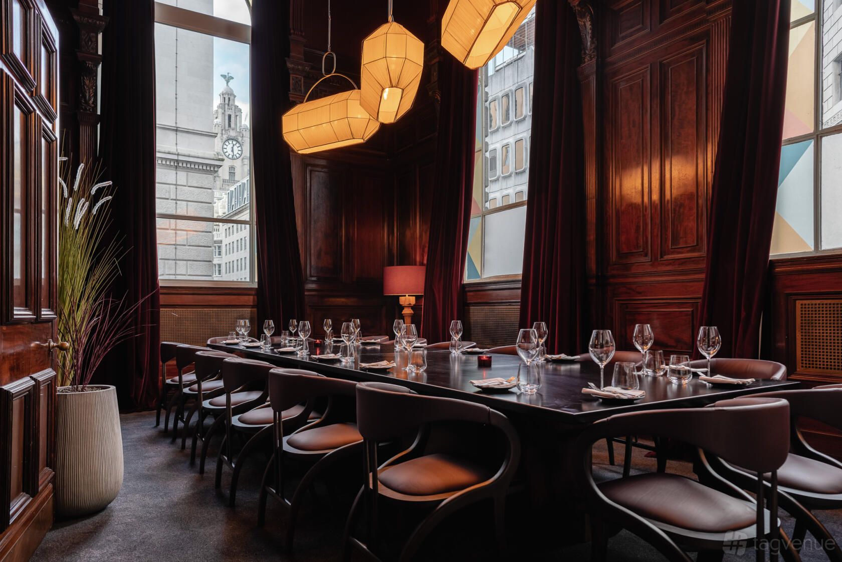 A dining room with dark wood paneling, pendant lighting, a long table set for a meal, and large windows at Gaucho Liverpool.