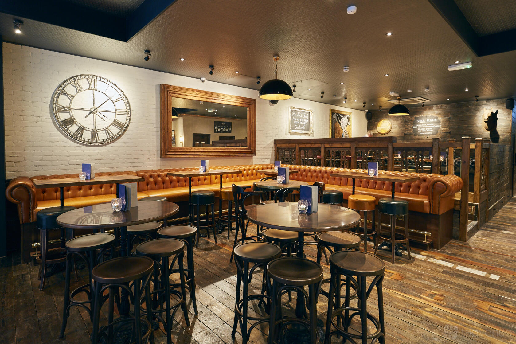 A pub interior with leather banquettes, round wooden tables, and a large wall clock at The Running Horse.