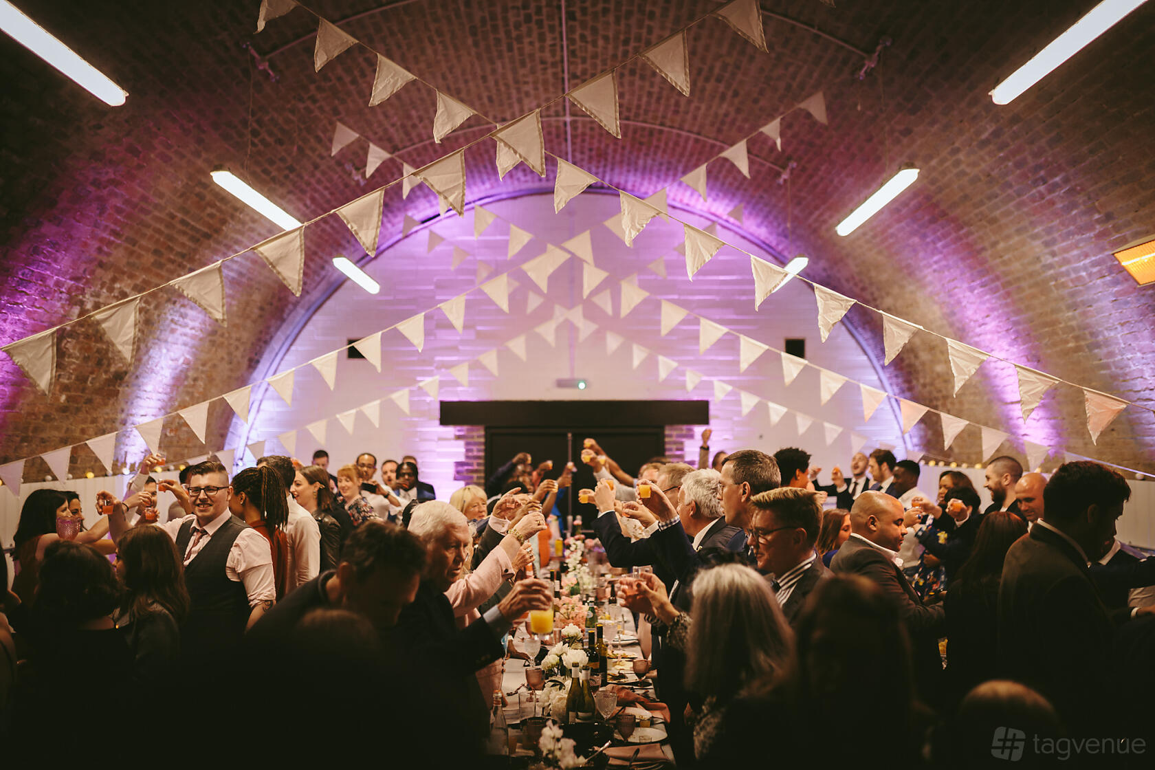 An event venue with arched brick ceilings, string bunting, and banquet tables under soft purple lighting at AMP Studios.