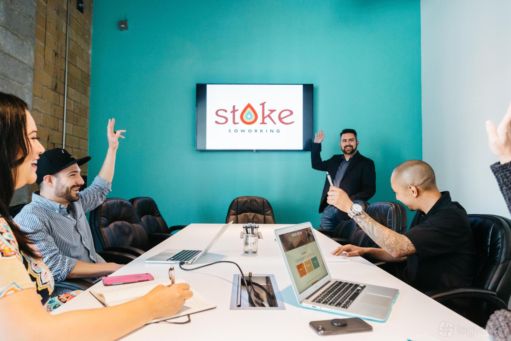 A coworking space with a large conference table, teal accent wall, and wall-mounted screen at Stoke Coworking.