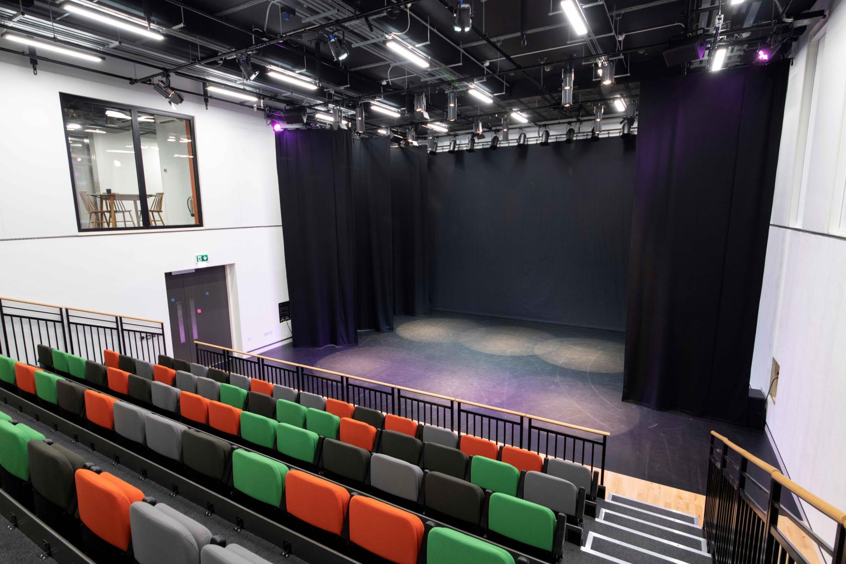 An event venue with tiered seating in orange, green, and gray facing a curtained stage and exposed lighting at The Dance Space.