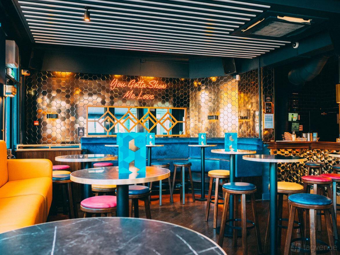 A bar with round tables, colorful stools, a blue velvet banquette, and a neon sign at Be At One Reading.