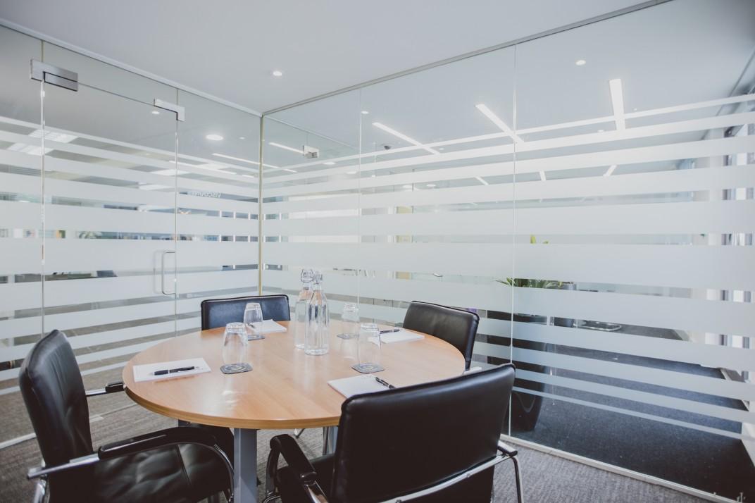 A meeting room with a round wooden table, black leather chairs, and frosted glass walls at Orega - The Beehive - Gatwick.