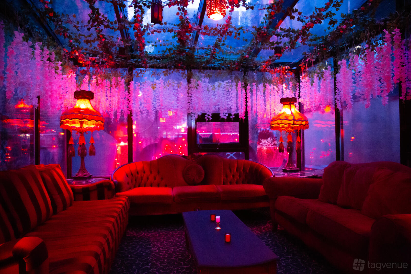 A lounge with red velvet sofas, vintage lamps, and hanging floral decor under a glass ceiling at Madame X.