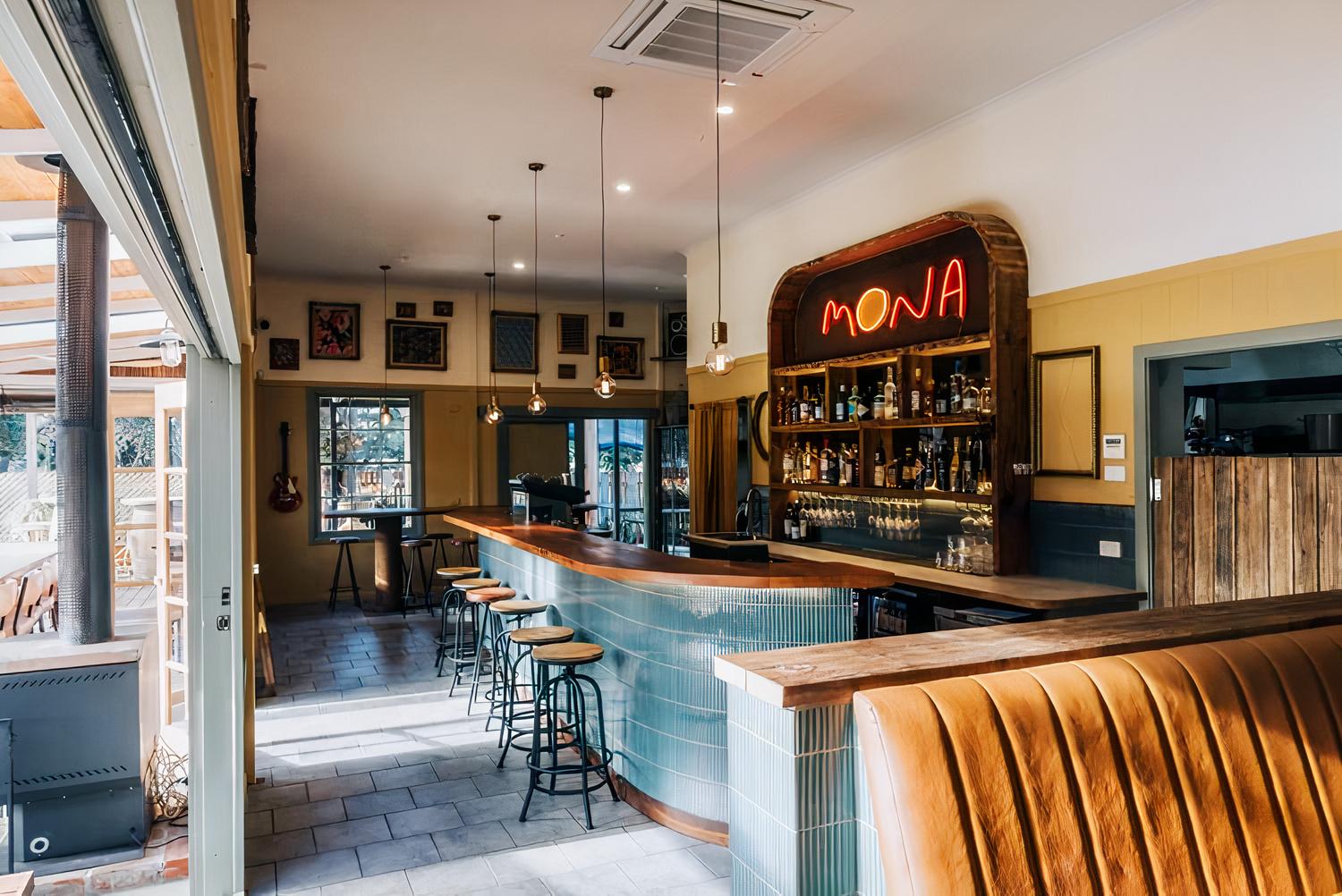 A restaurant with a curved tiled bar, backlit 'MONA' sign, and framed wall art at Mona Cicchetti Bar.