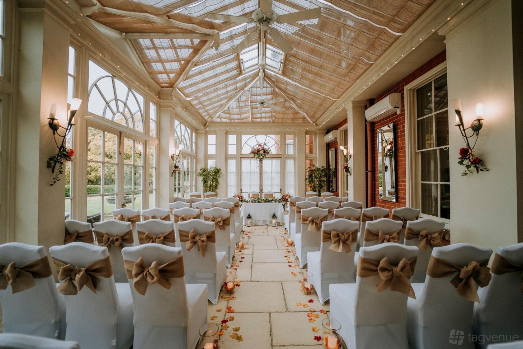 An event space with a glass ceiling, large arched windows, and rows of chairs with white covers and bows at Dovecliff Hall Hotel.