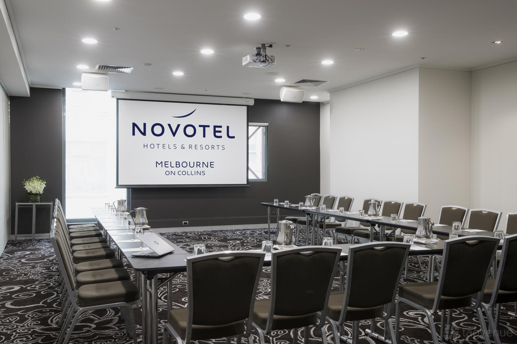 A function room with U-shaped tables, patterned carpet, and a projector screen at Novotel Melbourne on Collins.