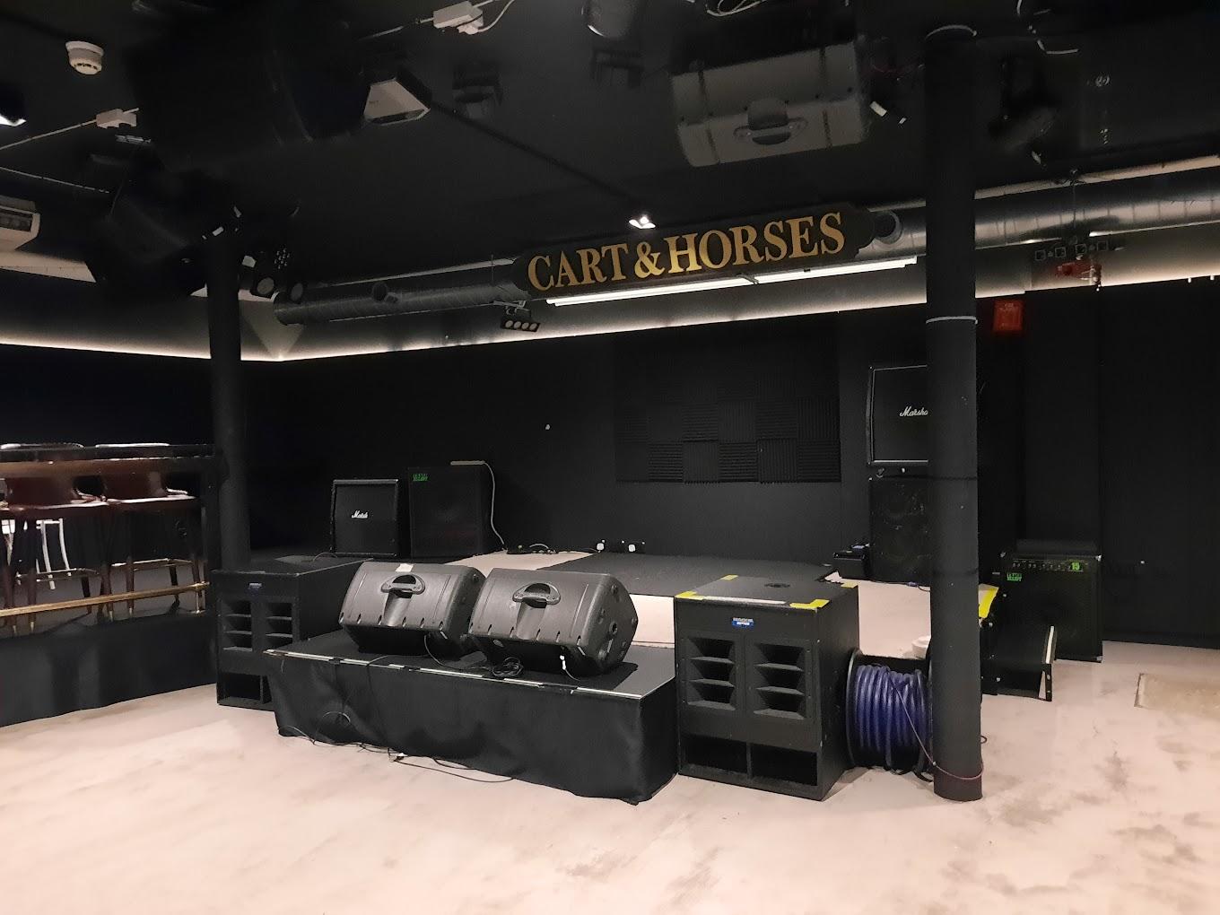 A basement venue with stage speakers, amplifiers, and a black backdrop beneath a Cart & Horses sign.