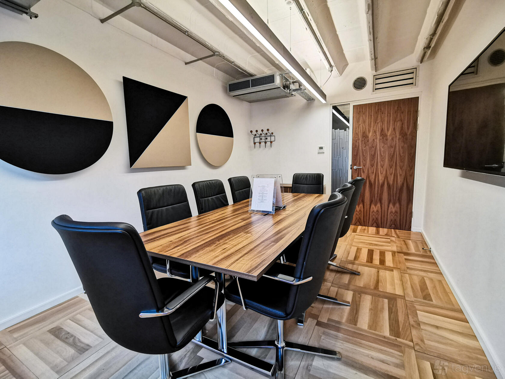 A boardroom in an office space with a wood meeting table, black leather chairs, abstract wall art, and parquet flooring at Us&Co Monument.