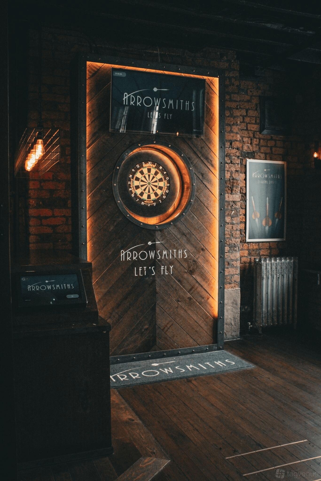 A pub event space with exposed brick walls, digital darts board, and wood paneling at Arrowsmiths @ The Shipping Forecast Liverpool.