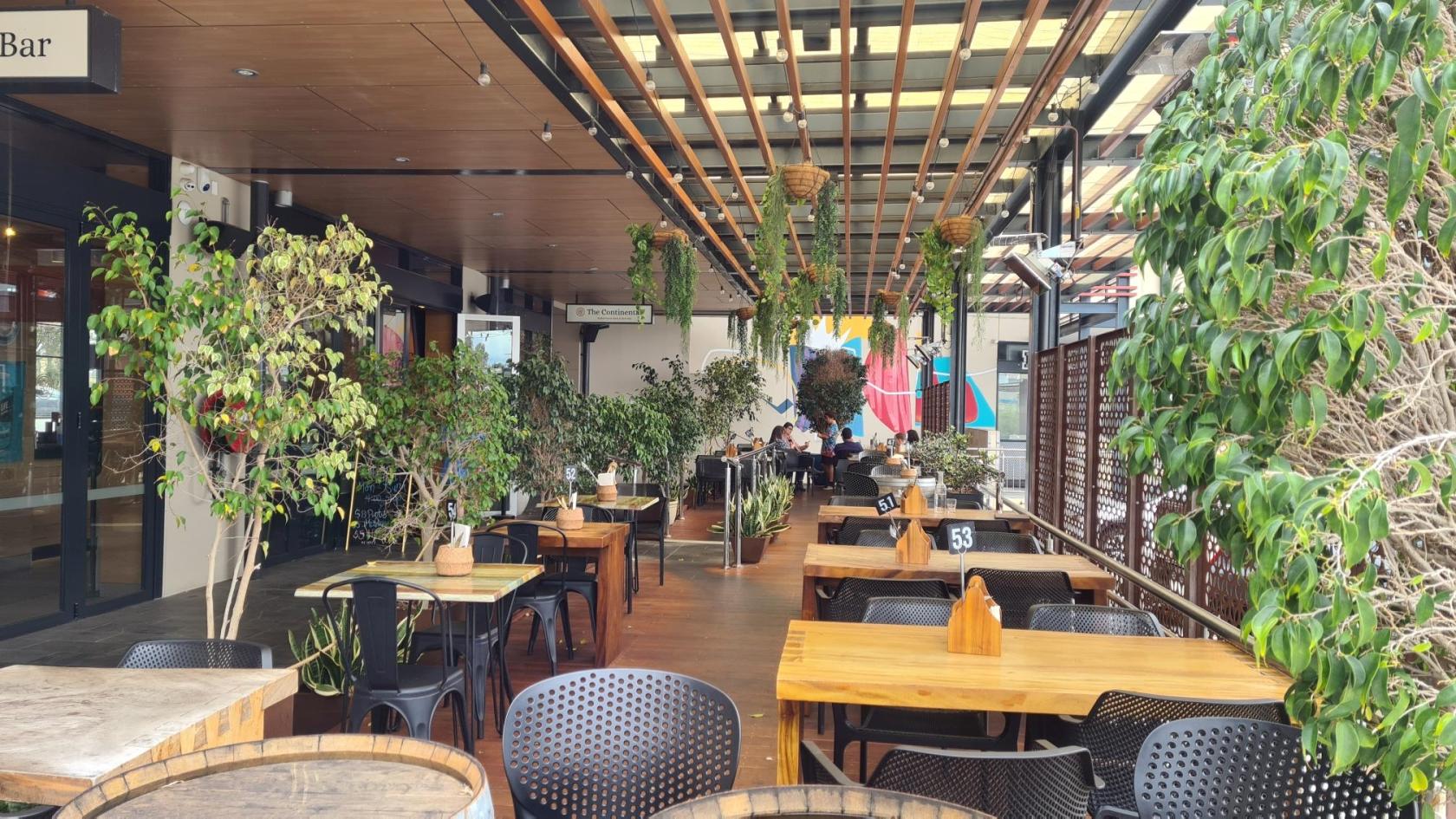 An outdoor dining area with wooden tables, hanging greenery, and potted plants at The Continental European Bar & Bistro.