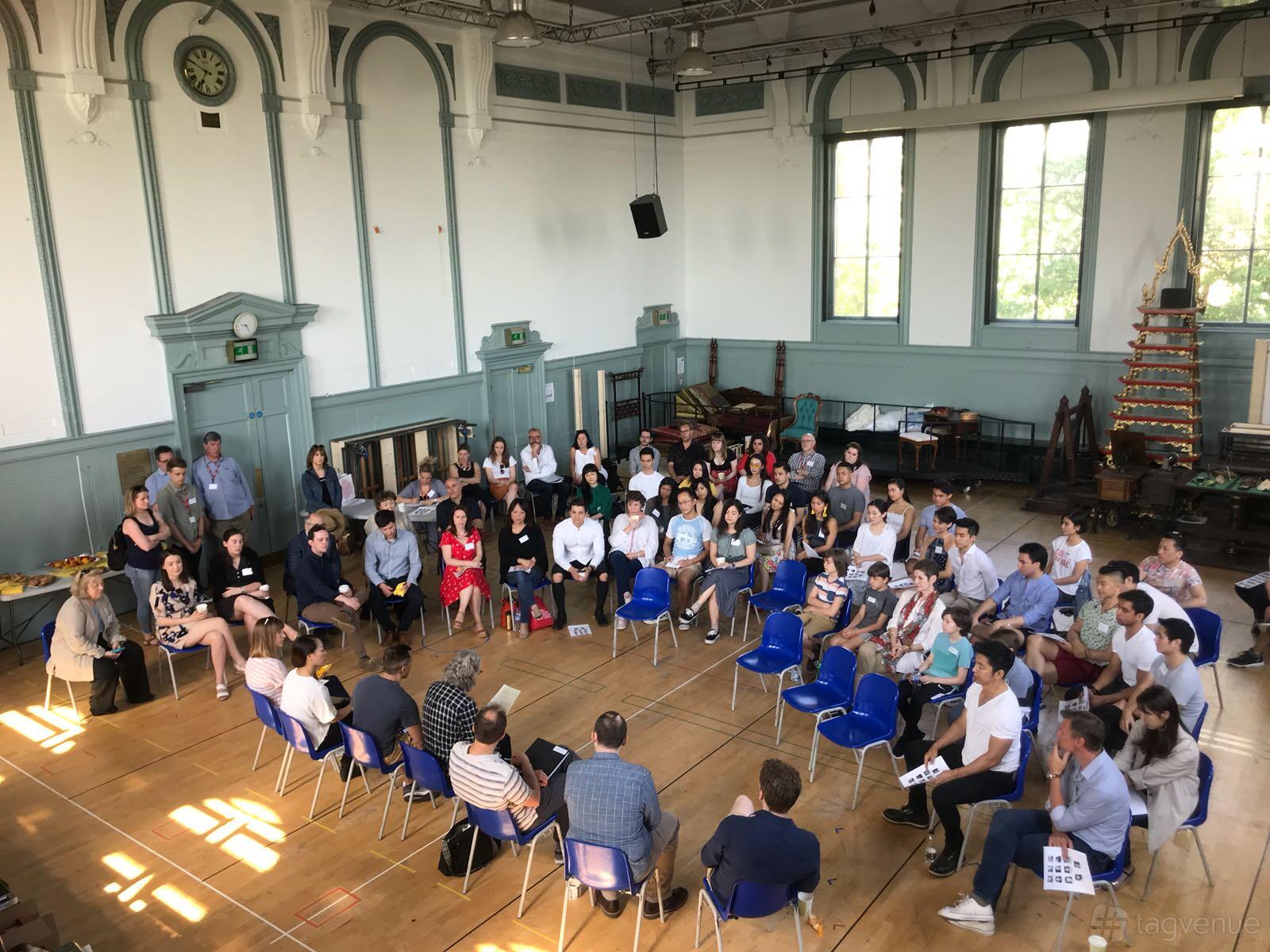 A spacious hall with high arched windows, wooden floors, and blue chairs arranged in a circle at Wac Arts.