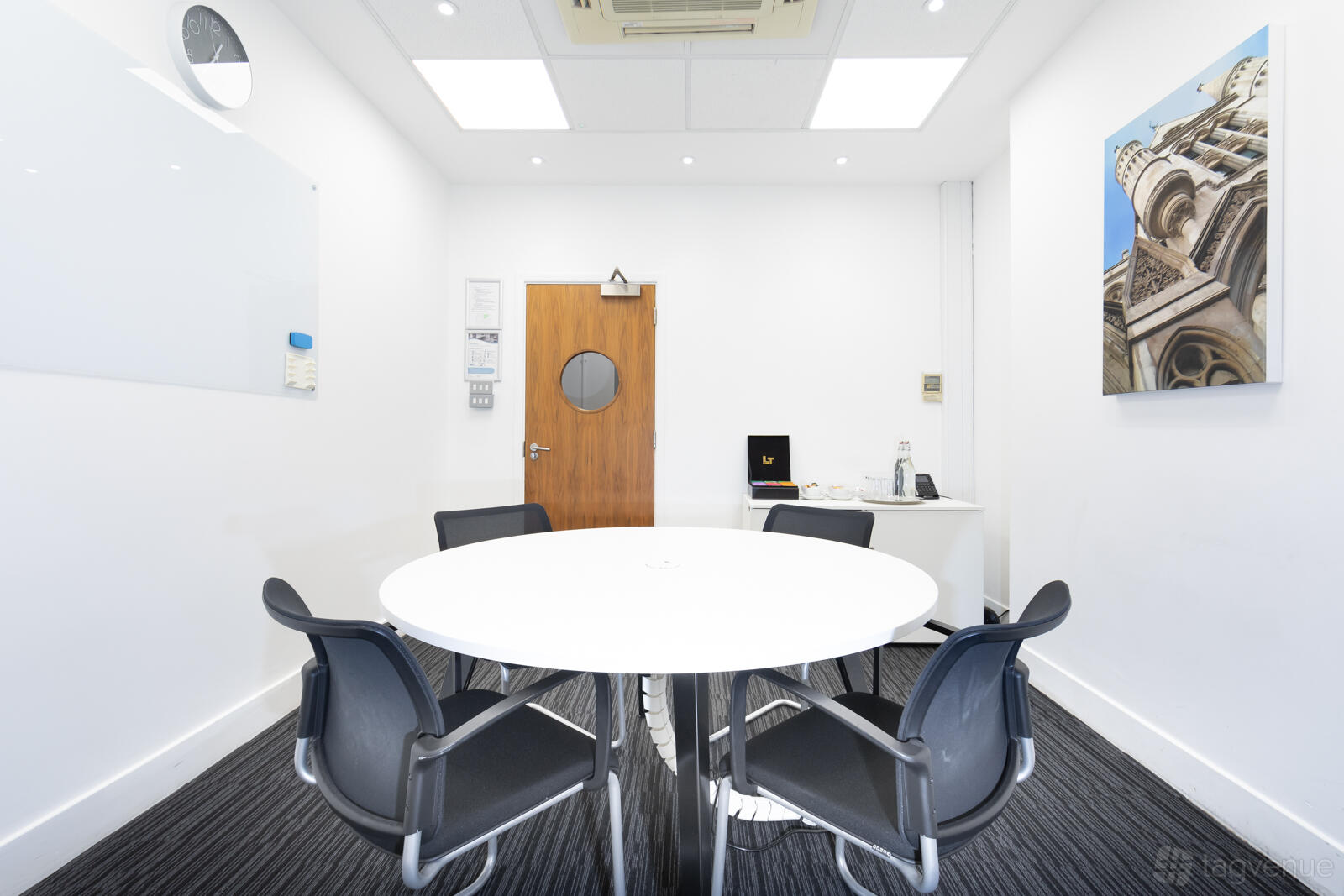 A meeting room with a round white table, five black chairs, wall art, and a whiteboard at 83 Victoria Street.