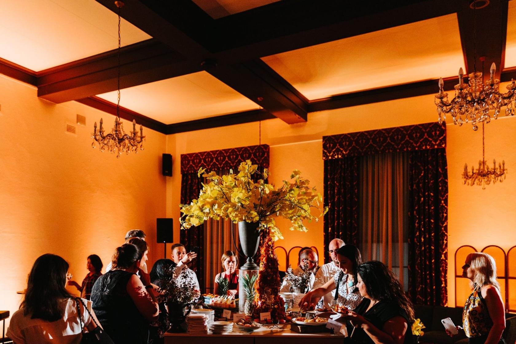An event venue with chandeliers, large windows draped with curtains, and a buffet table with a tall floral arrangement at HostCo Sydney Drawing Room.