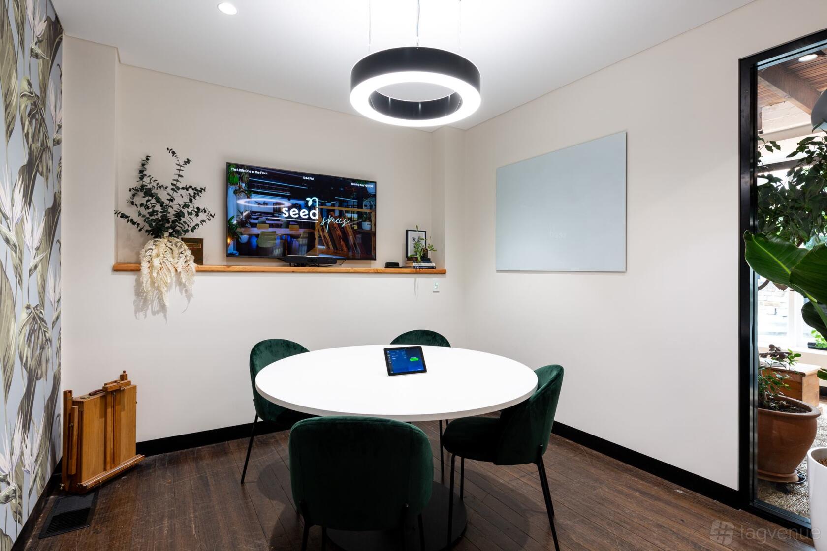 A meeting room with a round table, green velvet chairs, wall-mounted screen, and a whiteboard at Zeal Spaces.