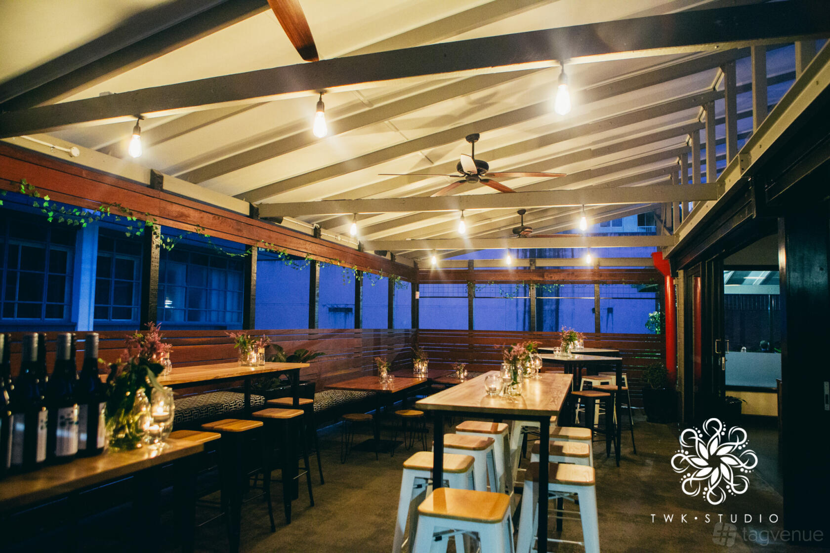 An event space in a pub with long wooden tables, stools, ceiling fans, and string lights at The Transcontinental Hotel.