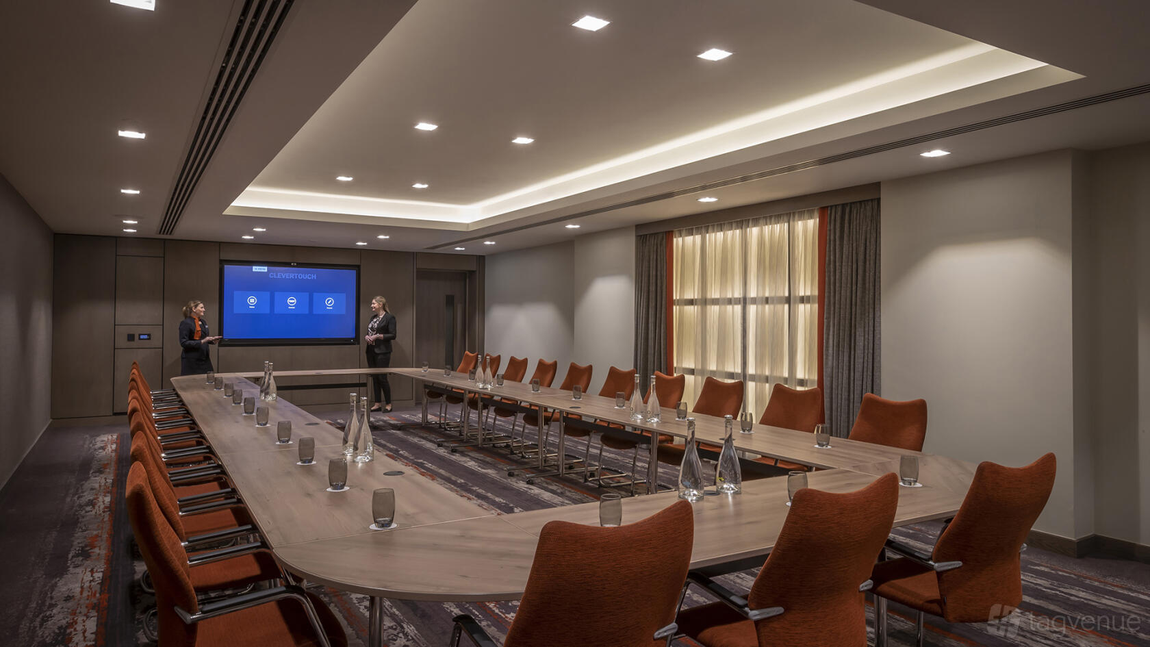 A hotel meeting room with U-shaped wood tables, orange chairs, and a large presentation screen at Clayton Hotel Cardiff Lane.
